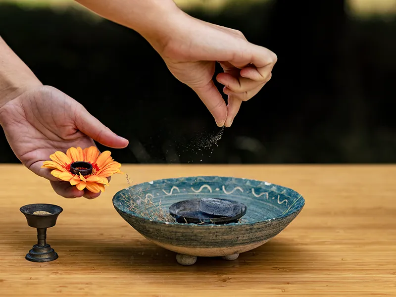 Op een houten tafel staat links een kelk waar wierrookpoeder in zit. Rechts daarvan staat een stenen pot met daarin een kooltje. Een hand houdt een bloem vast met daarin een ring. Met het andere hand wordt er wierrook gestrooid op het kooltje.