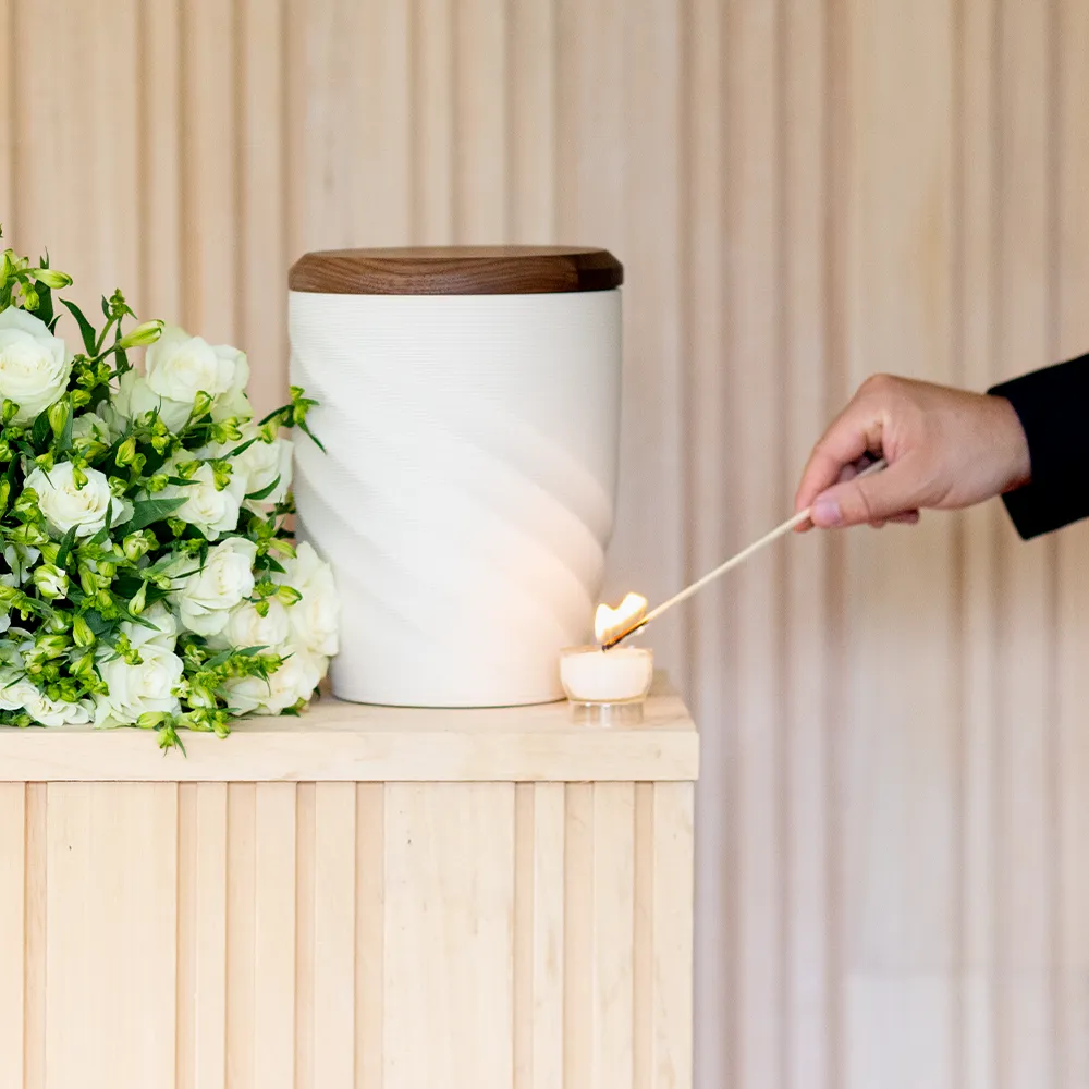 Er staat een witte urne met een rouwboeket op een houten hoge tafel. De hand van Ruben steekt een wit theelichtje aan.