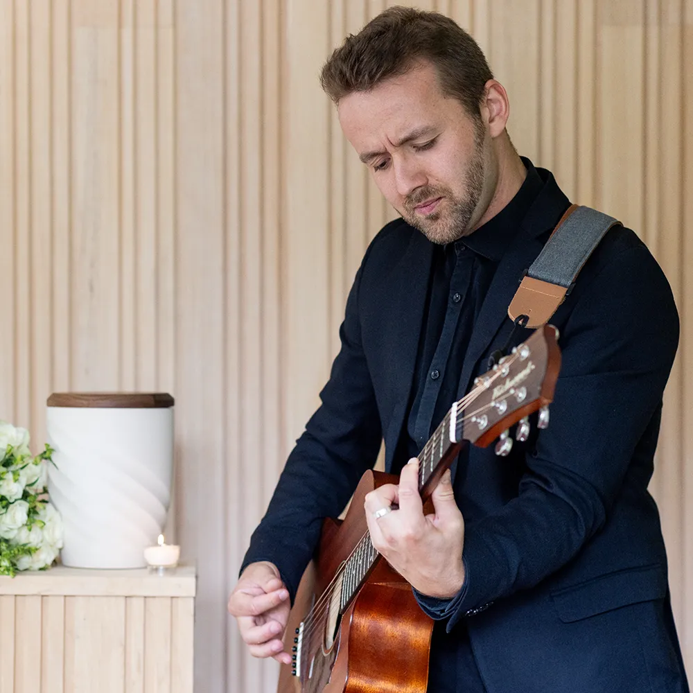 Ruben staat naast een urne met een rouwboeket en een wit theelichtje. zijn ogen zijn gesloten en hij speelt gitaar.