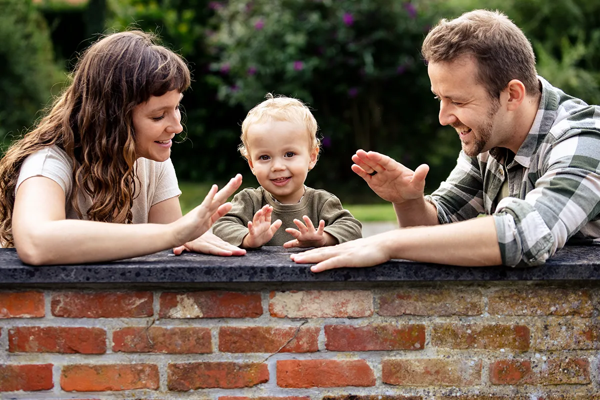 Ruben, Elien en Eliah staan bij een laag muurtje. Ze slaan met hun handen op de muur als een spelletje. Elien en Ruben kijken naar Eliah. En Eliah kijkt in de lens. Ze genieten van het samen spelen.