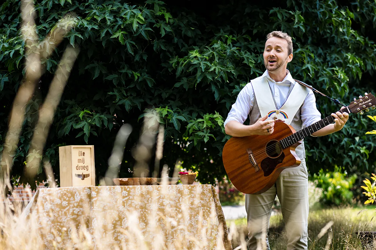 Ruben staat rechts van een tafel met zijn gitaar. Hij heeft een kostuum aan en een doopkraag. De tafel staat buiten bij een boom. Op de tafel staat een kaars, een doopschaal en enkele bloemen.
