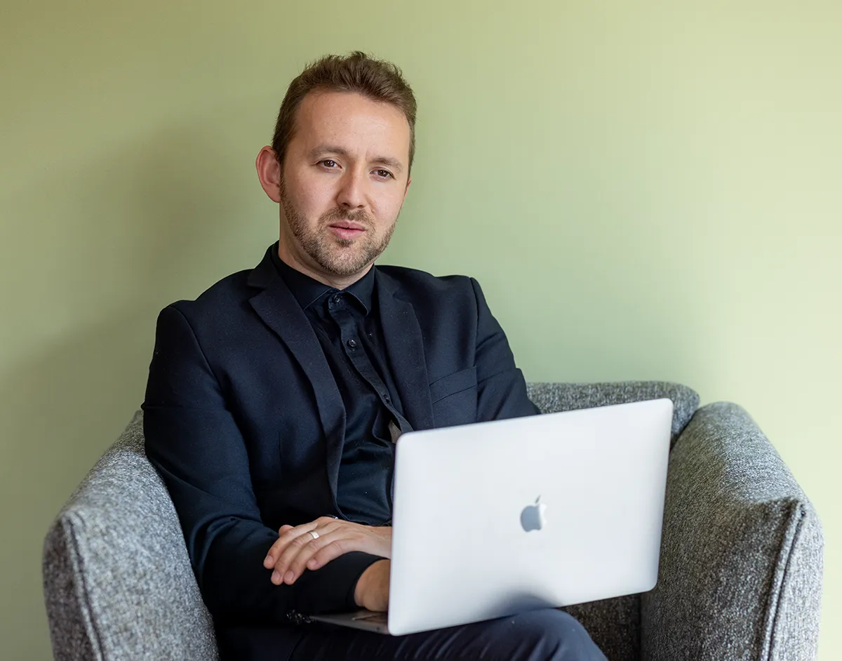 ruben zit in een zetel. Hij kijkt met een blik vol compassie naar iemand waar hij tegen aan het praten is. Hij heeft een zwart kostuum aan en een laptop op zijn schoot.