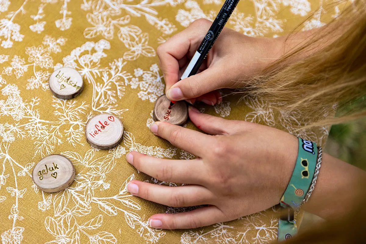 Jonge handen schrijven op houten ronde schijfjes spreuken om bij stil te staan zoals blijdschap, liefde, geluk,...