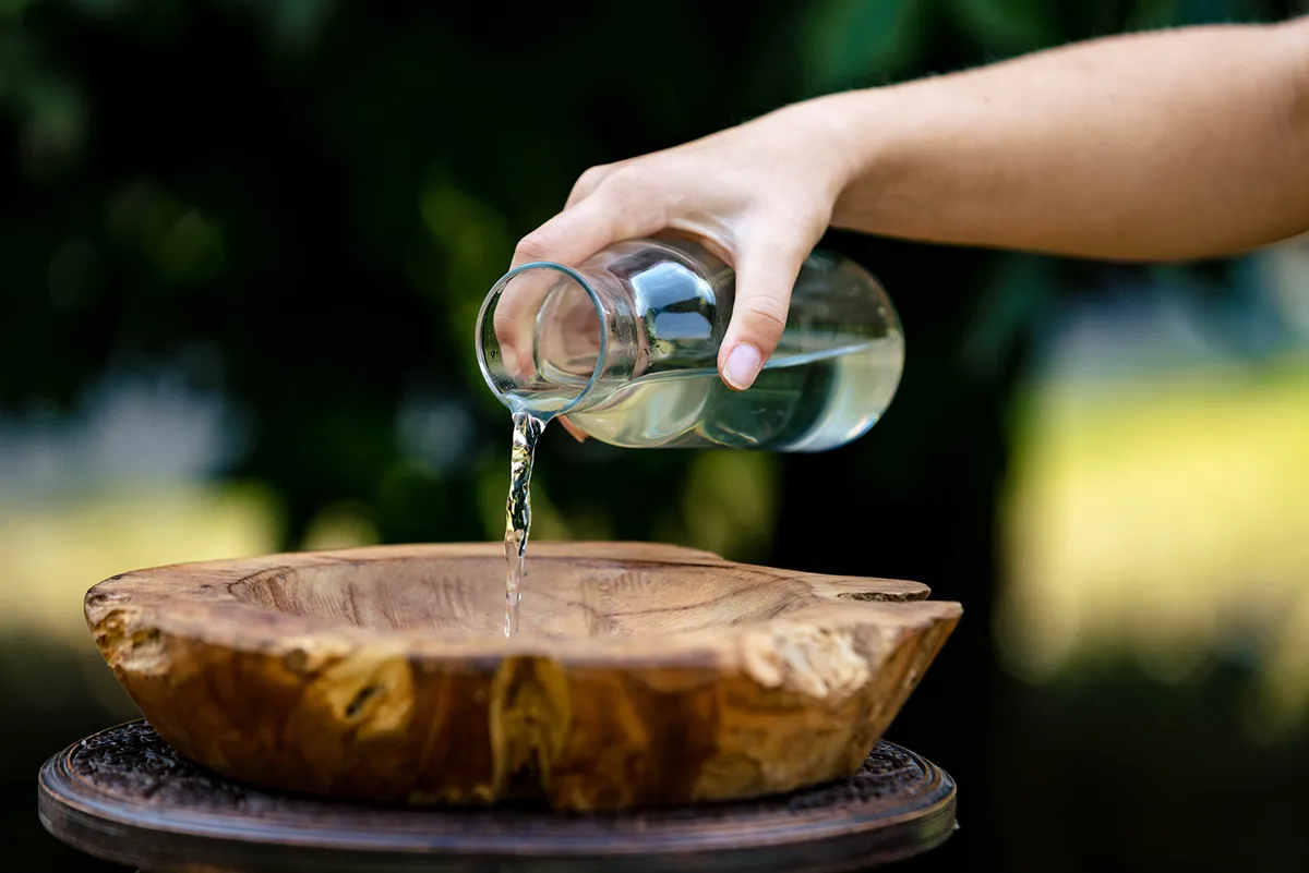 De houten doopschaal op het houten tafeltje staat in de natuur staan. Een hand giet water uit een glazen fles in de doopschaal.