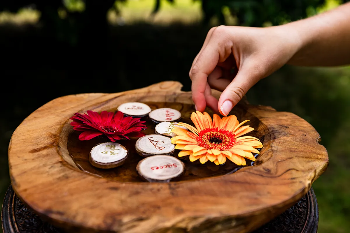 Er staat een houten doopschaal op een houten tafeltje buiten in de natuur. In de doopschaal zit water, bloemen en houten, ronde schijfjes met spreuken die worden meegegeven. Een hand legt de bloem in de doopschaal.